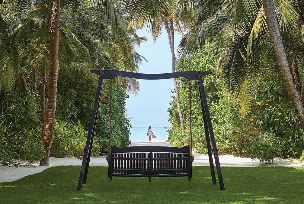 A swing surrounded by trees looking out an opening at a woman in the distance on the beach