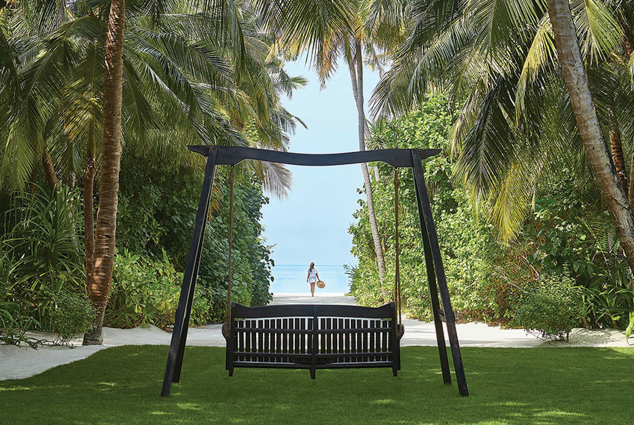A swing surrounded by trees looking out an opening at a woman in the distance on the beach