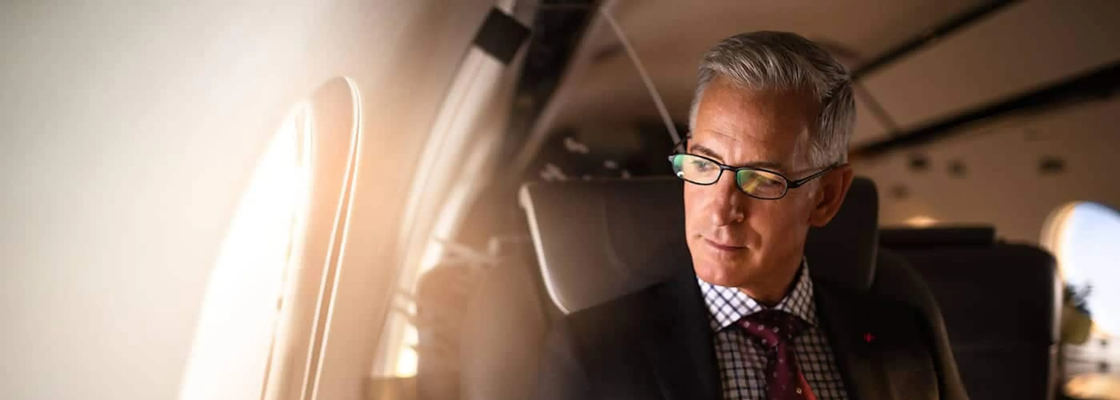 A businessman looks out a window of a private jet, seated with pen in hand and notebook