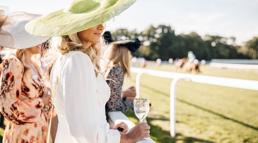 a lady in a big green hat smiles and looks on at the horse racing