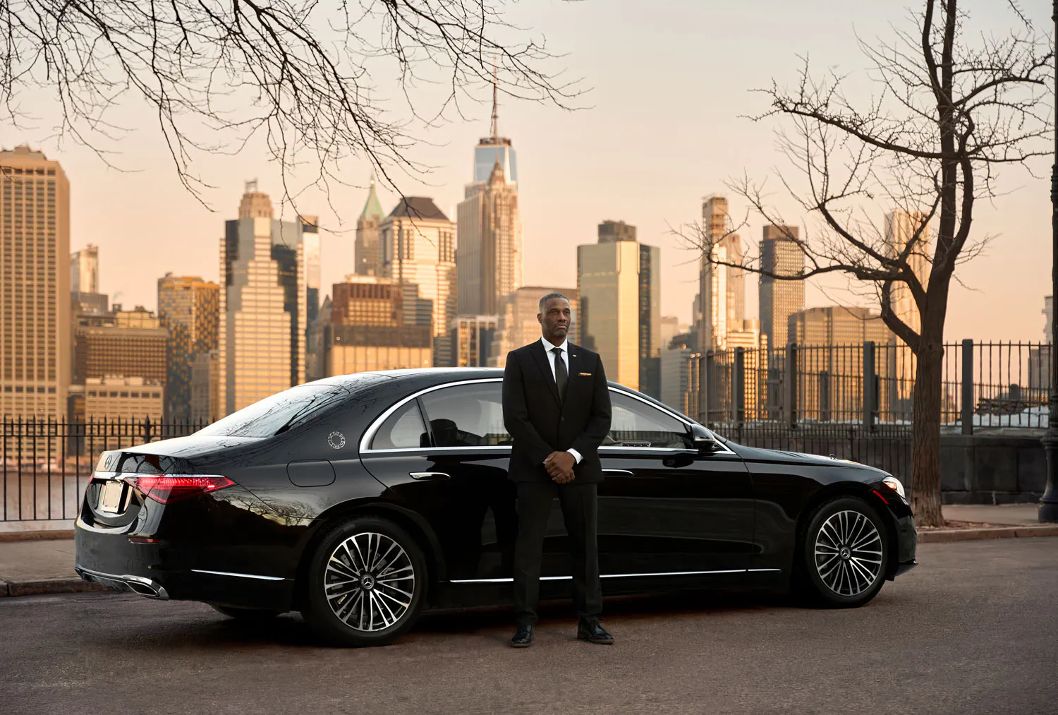 A man in suit and hands folded stands waiting outside a Wheely branded black car