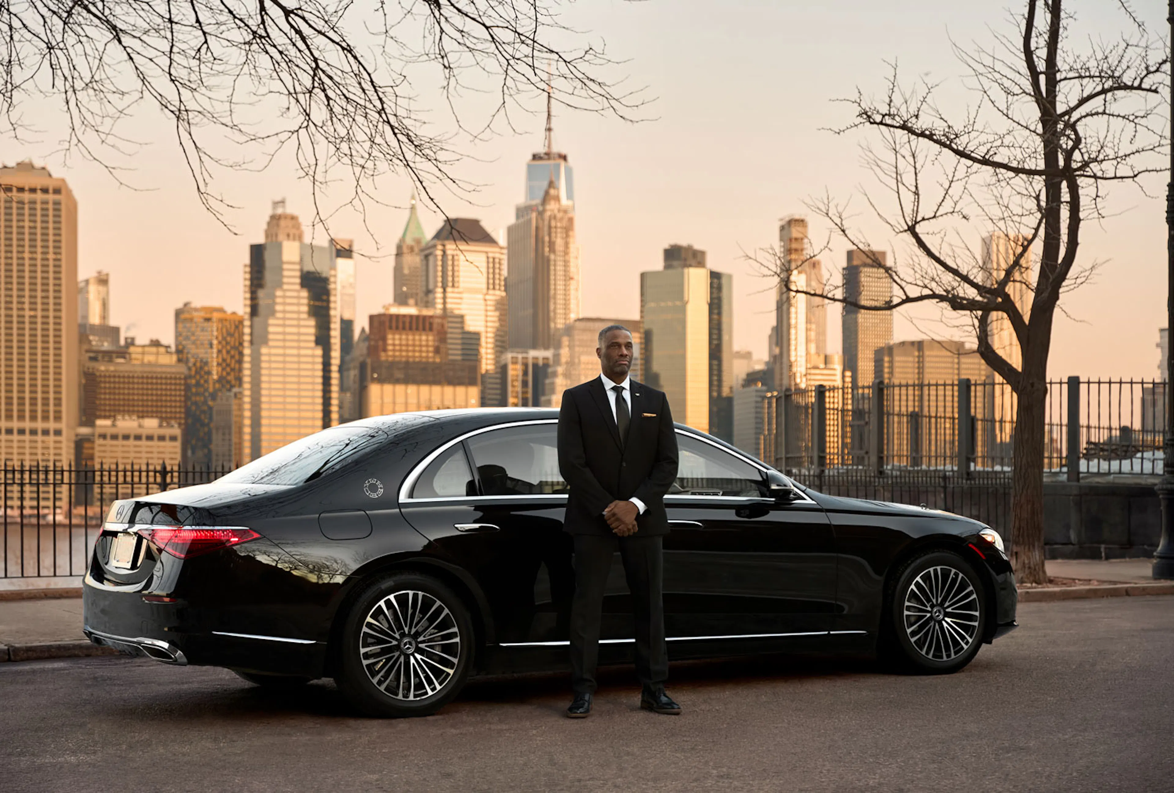 A man in suit and hands folded stands waiting outside a Wheely branded black car 
