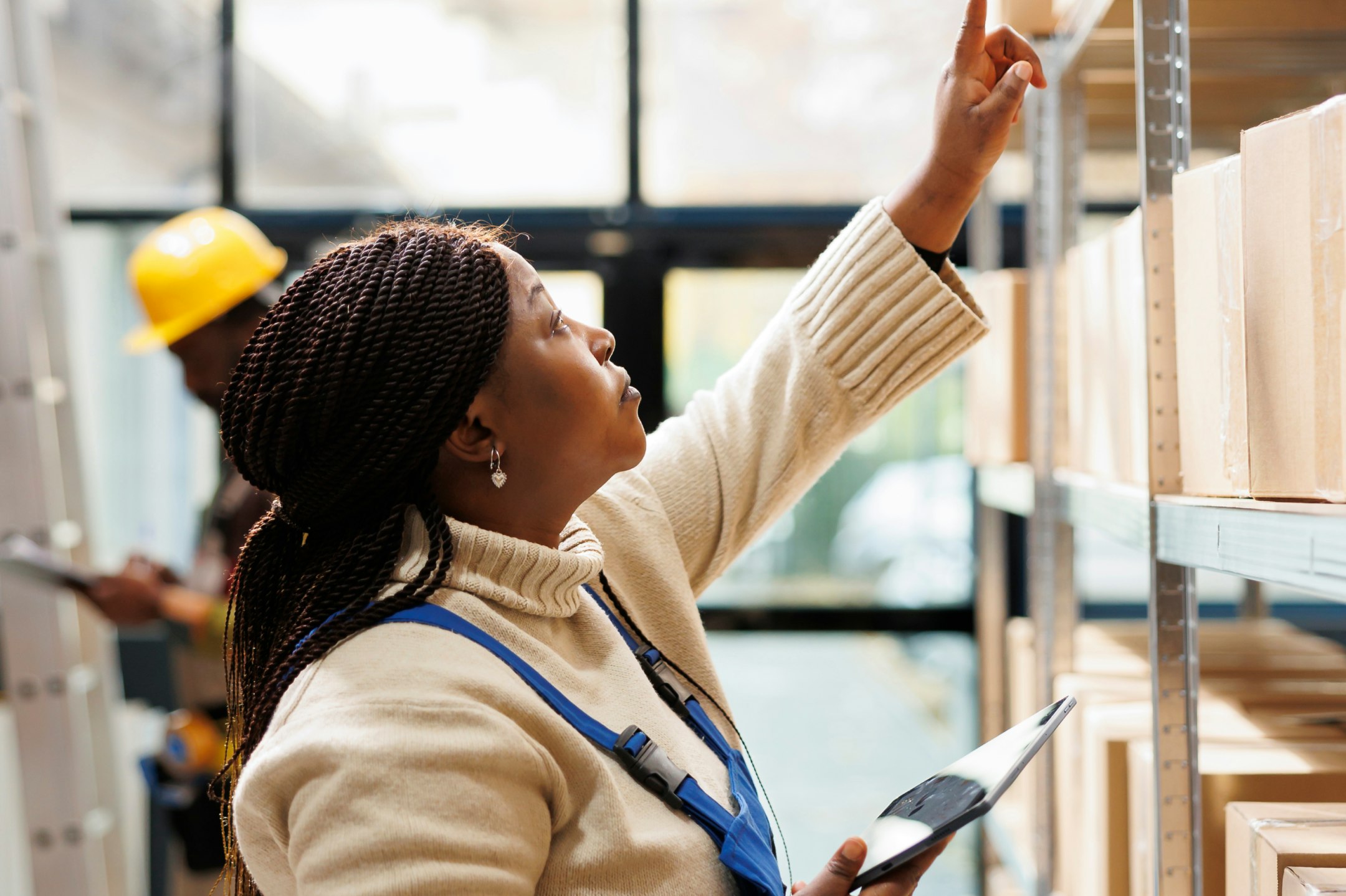 Frontline worker with long dark box braids in overalls checking warehouse shelves while holding a tablet