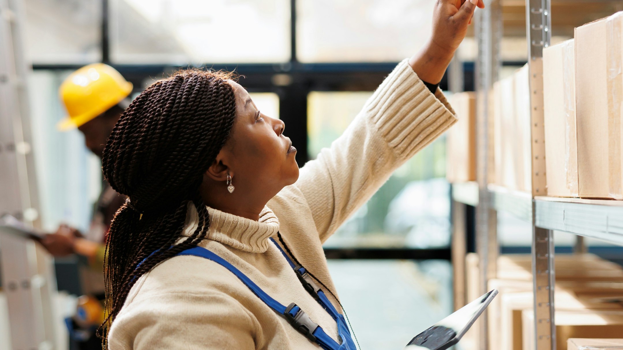 Frontline worker with long dark box braids in overalls checking warehouse shelves while holding a tablet