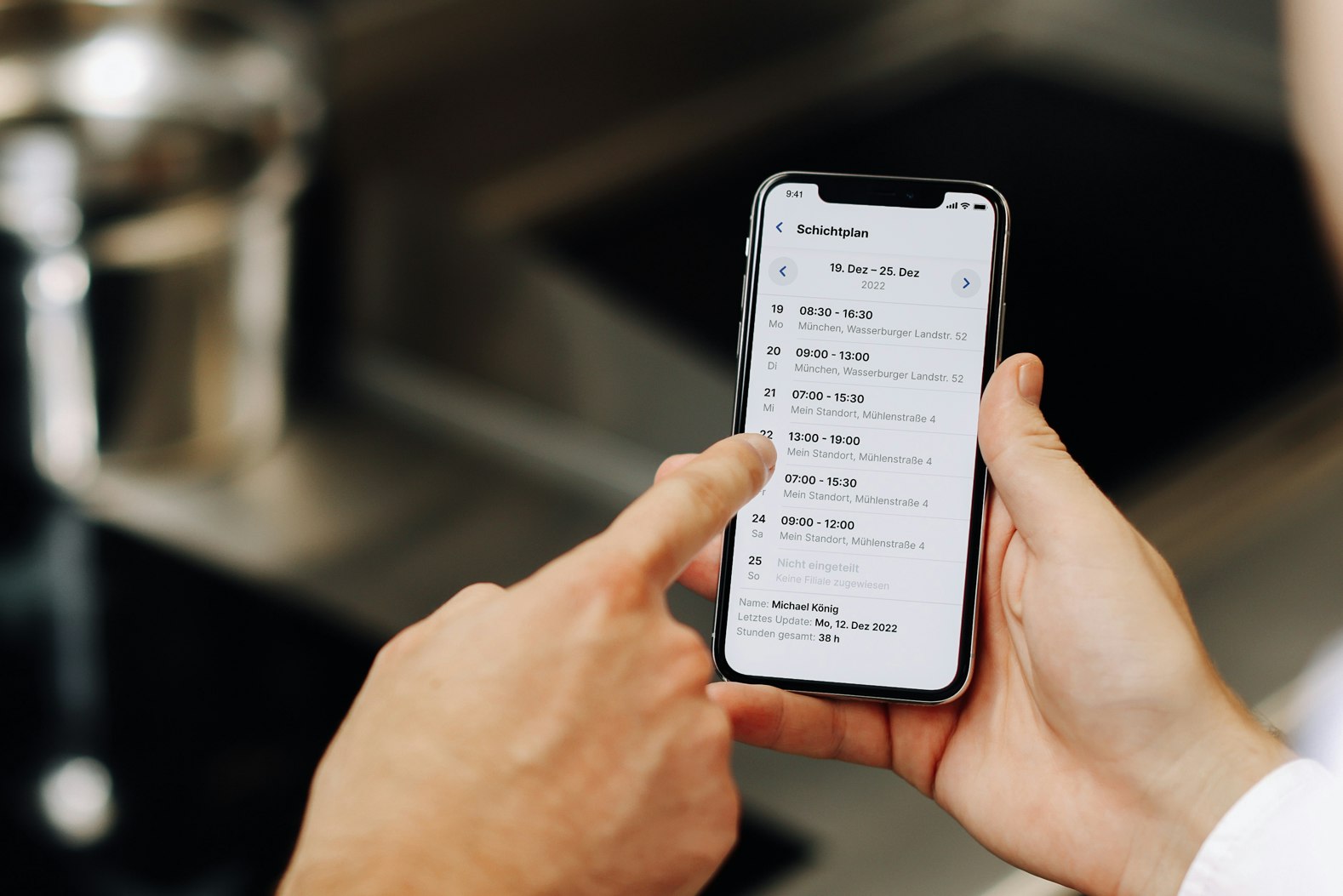 Smartphone-Display zeigt Schichtplan und Industrieküche im Hintergrund