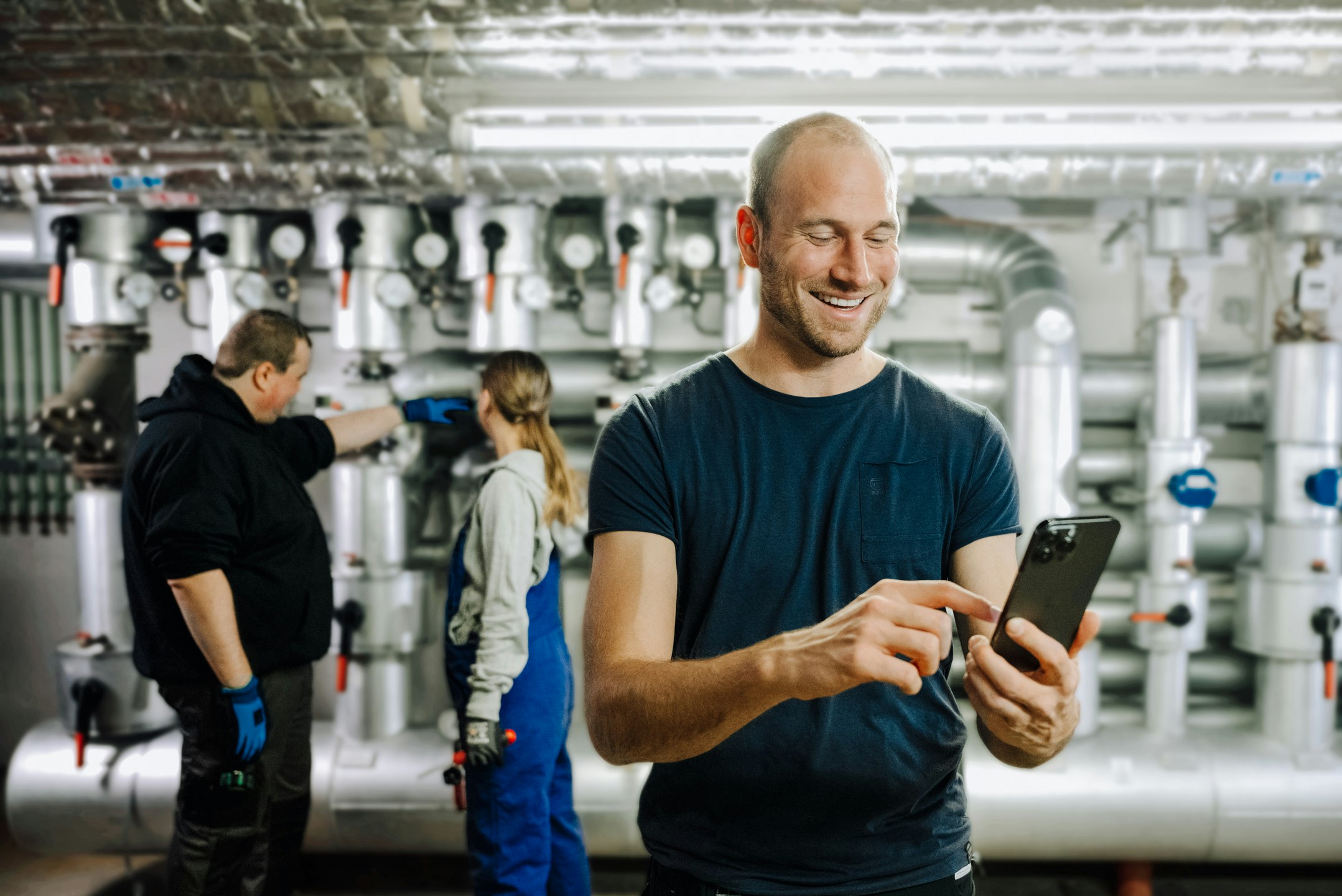 Worker operates smartphone while colleagues maintain industry equipment