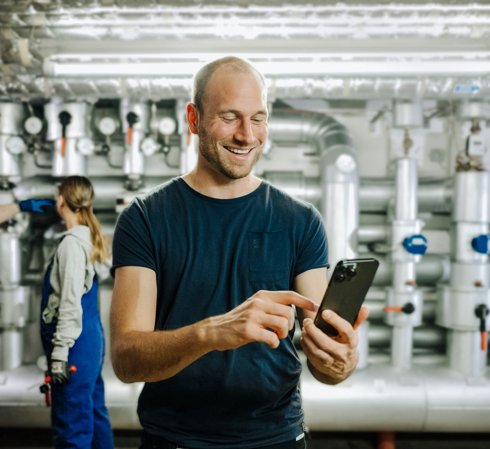 Worker operates smartphone while colleagues maintain industry equipment