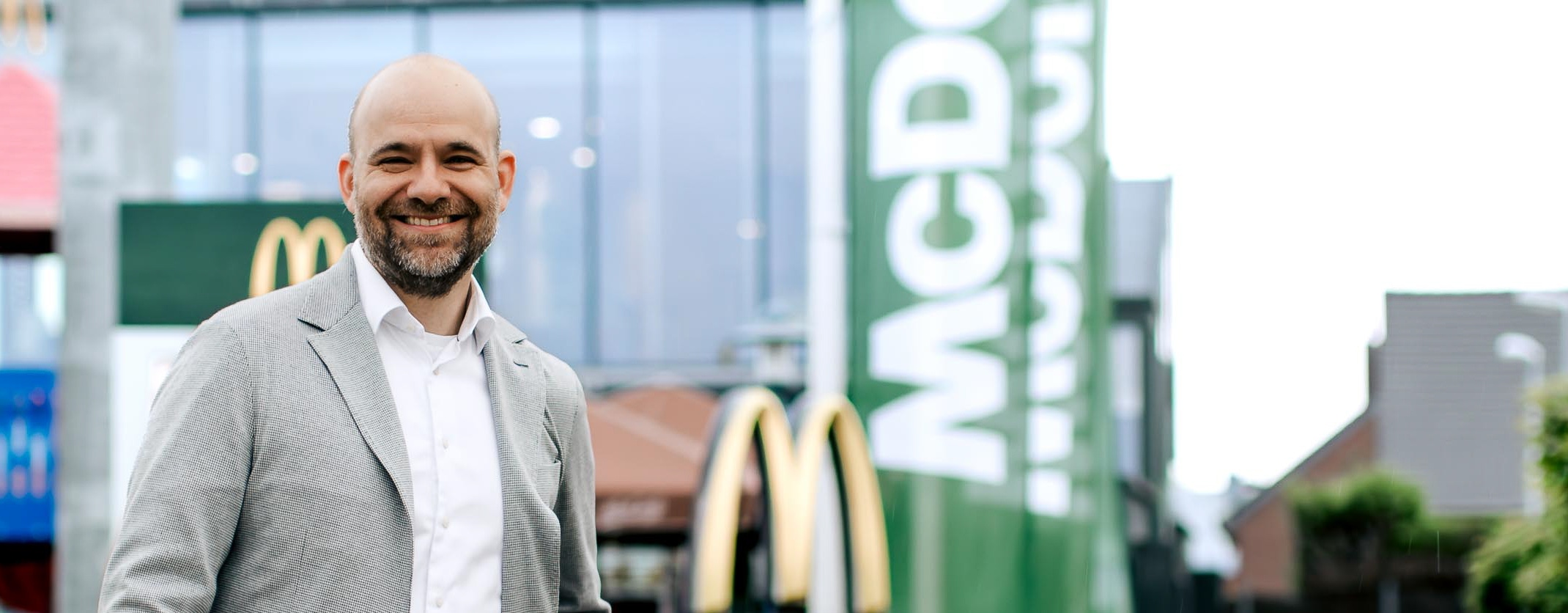 Mann lächelt in die Kamera. Er trägt eine graue Jacke und steht vor einem McDonald's Restaurant.
