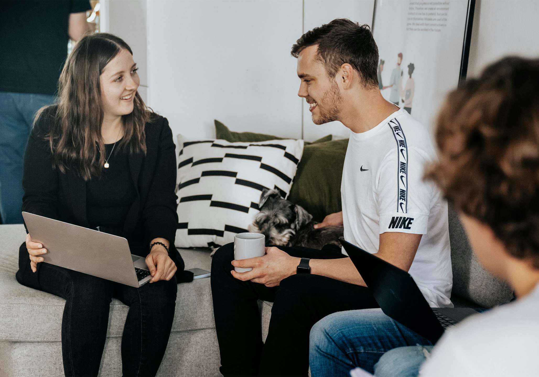 Junge Frau sitzt mit Kollegen und Bürohund auf dem Sofa 