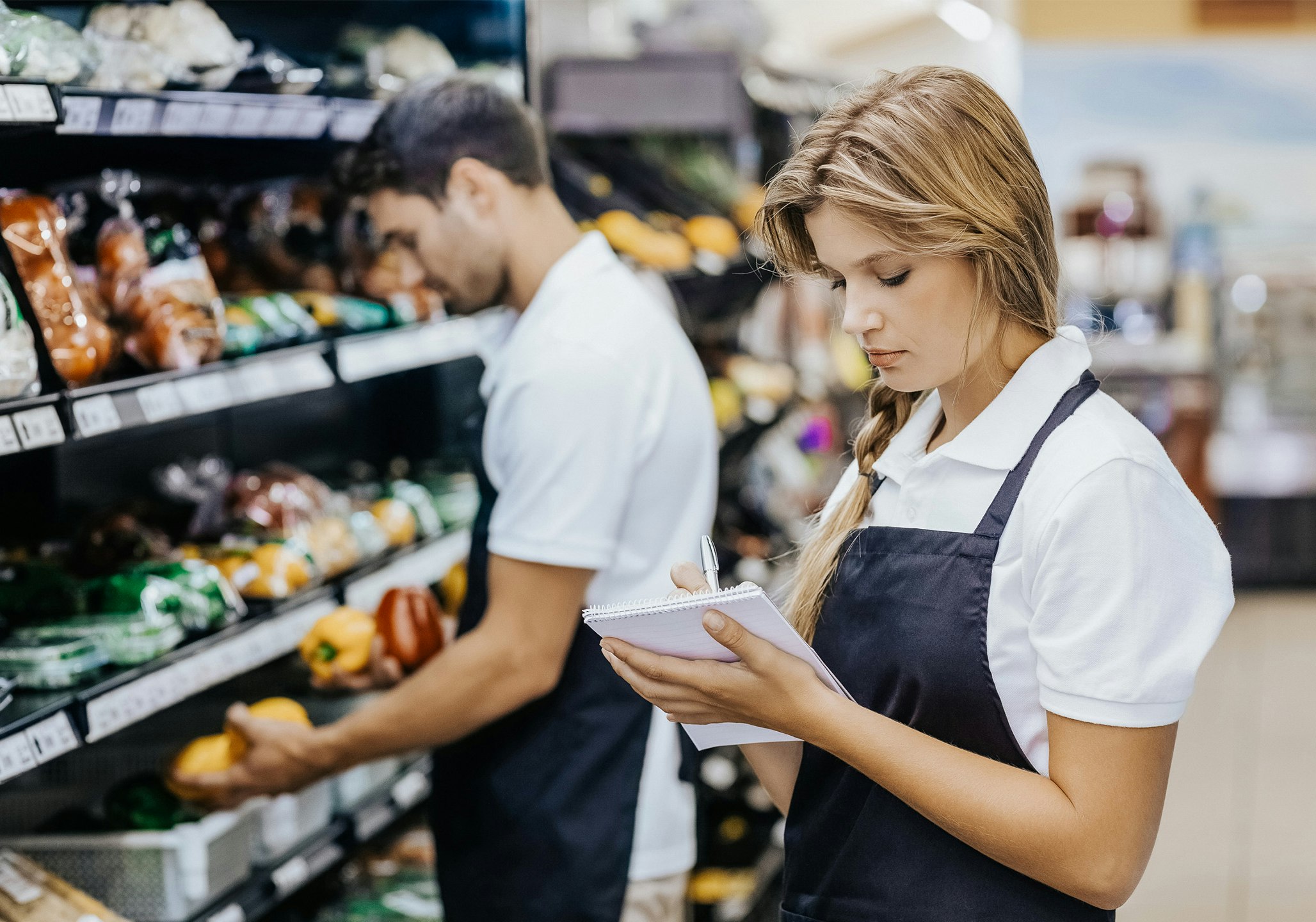 Frau steht vor Supermarktregal mit Schreibblock in der Hand und Mitarbeiter im Hintergrund räumt Regal ein