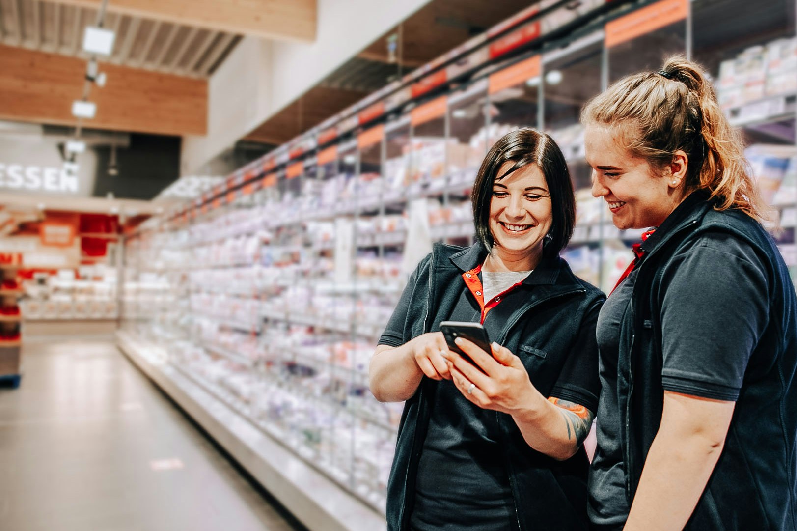 Zwei weibliche REWE Angestellte schauen auf ein Mobiltelefon