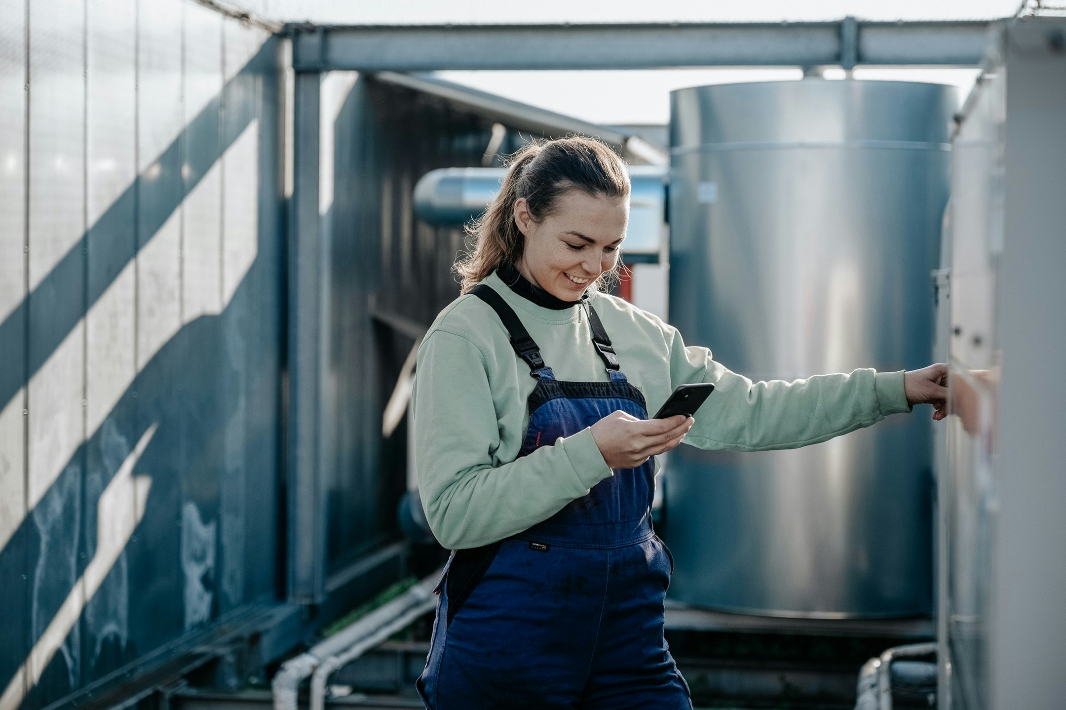 Lächelnde Frau im Blaumann mit Smartphone in der Hand vor Industrieanlage