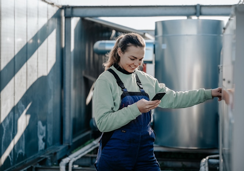 Lächelnde Frau im Blaumann mit Smartphone in der Hand vor Industrieanlage