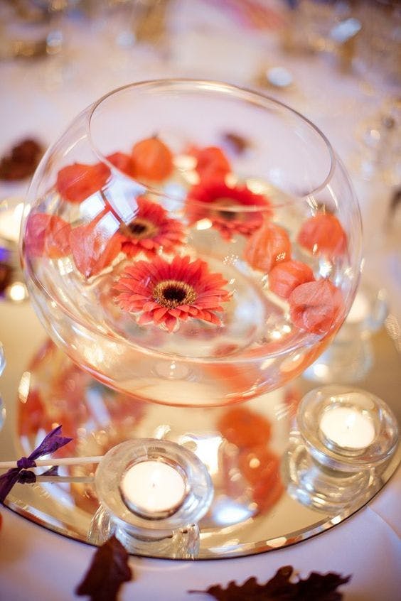 Single red gerberas and petals in fishbowls filled with water as centrepeices, surrounded by tea light candles and oak leaves scattered on table.