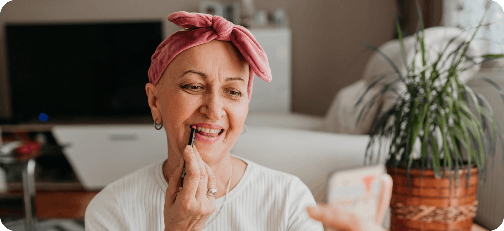 An older woman in a headscarf applies makeup while experiencing the effects of chemo and other cancer treatments on her hair.