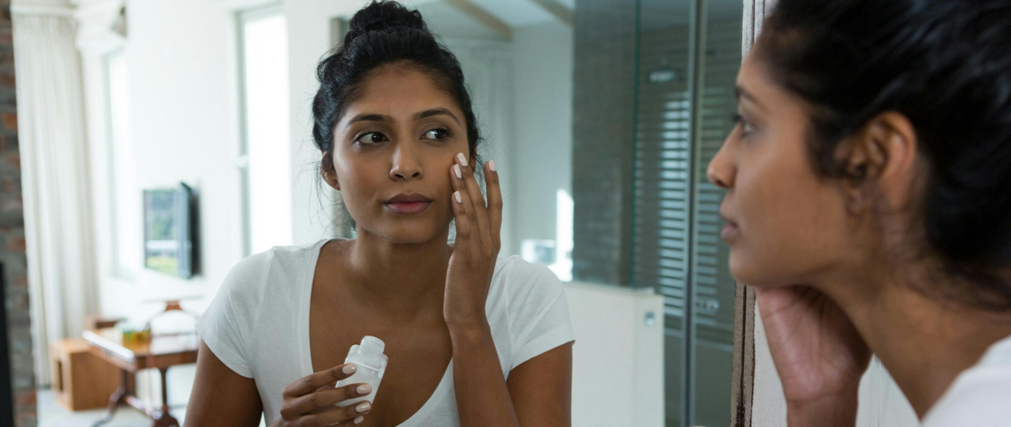 A woman standing in front of a mirror, looking at her reflection as she does her skincare routine.
