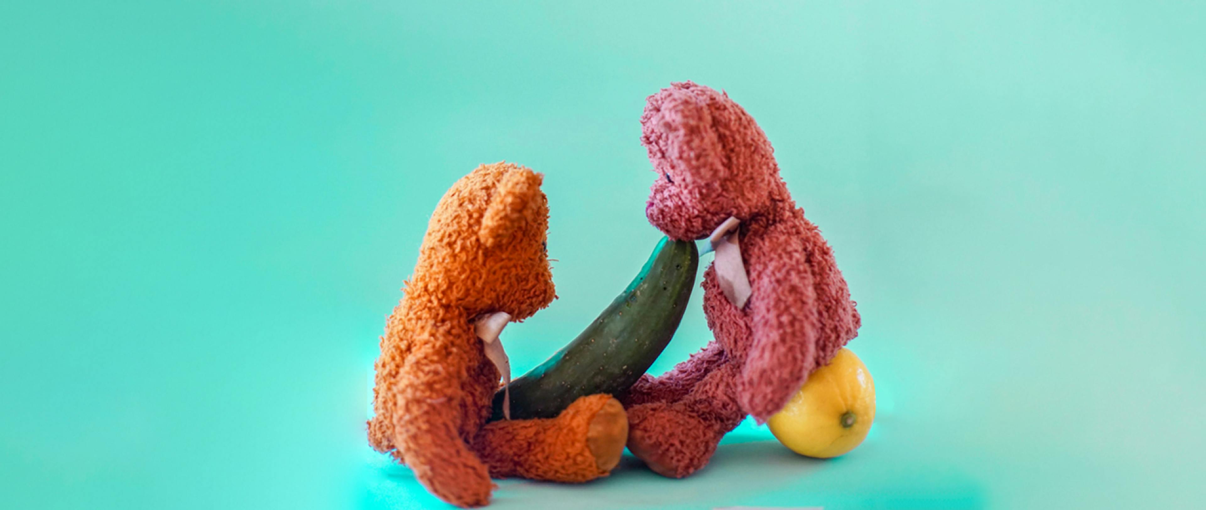 A pink and orange teddy bear with a cucumber between them meant to represent oral sex