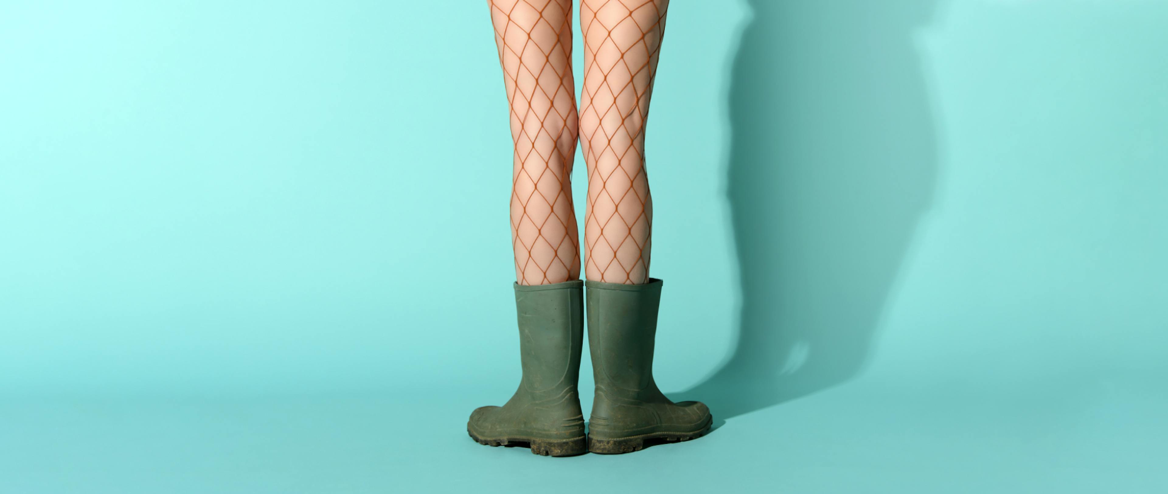 Crop unrecognizable female model wearing fishnet stockings and dirty rubber boots standing in blue studio.