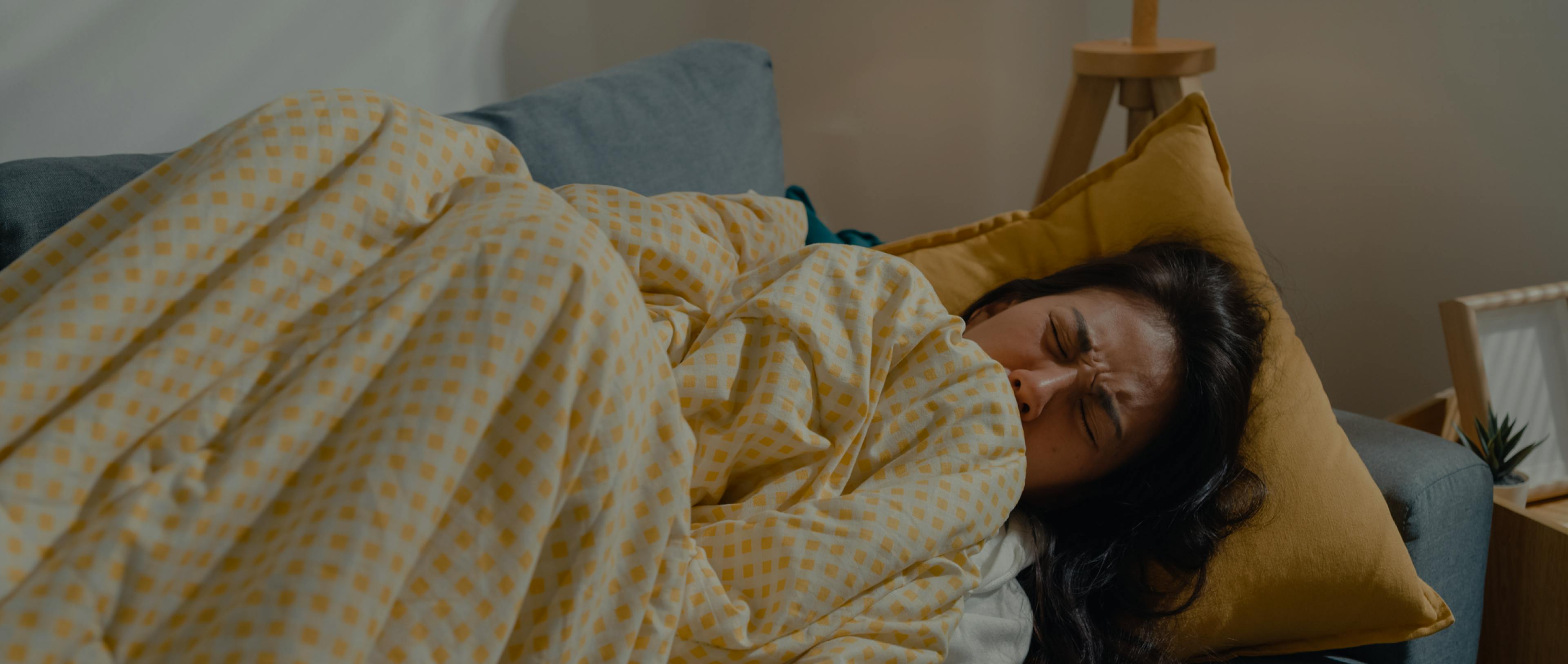 A sick young Asian woman lying down under the blanket on the sofa in the living room at home.