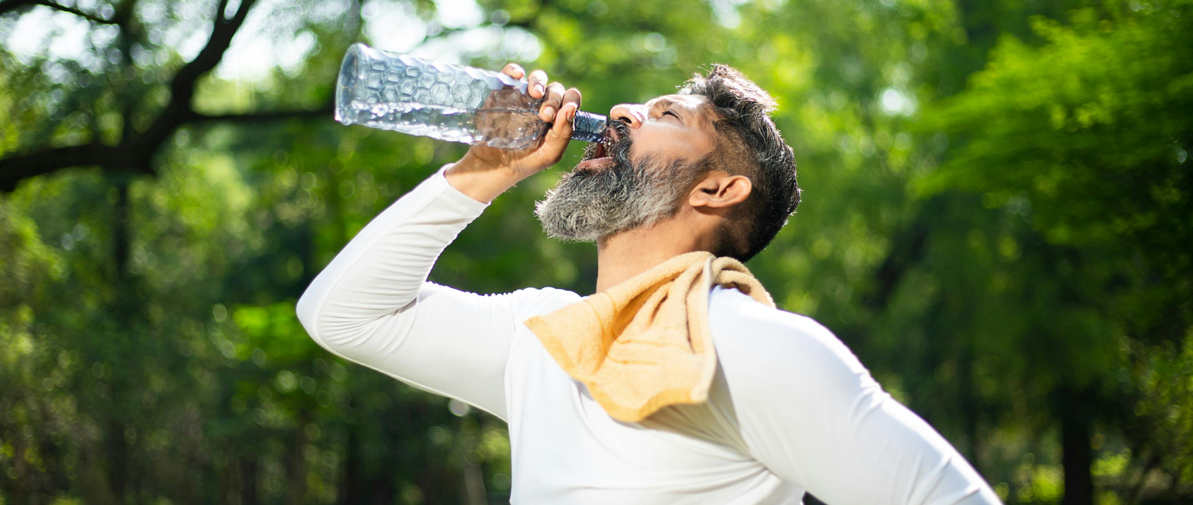 A tired and thirsty Asian man takes a break while drinking water from a bottle in the park on a hot day.