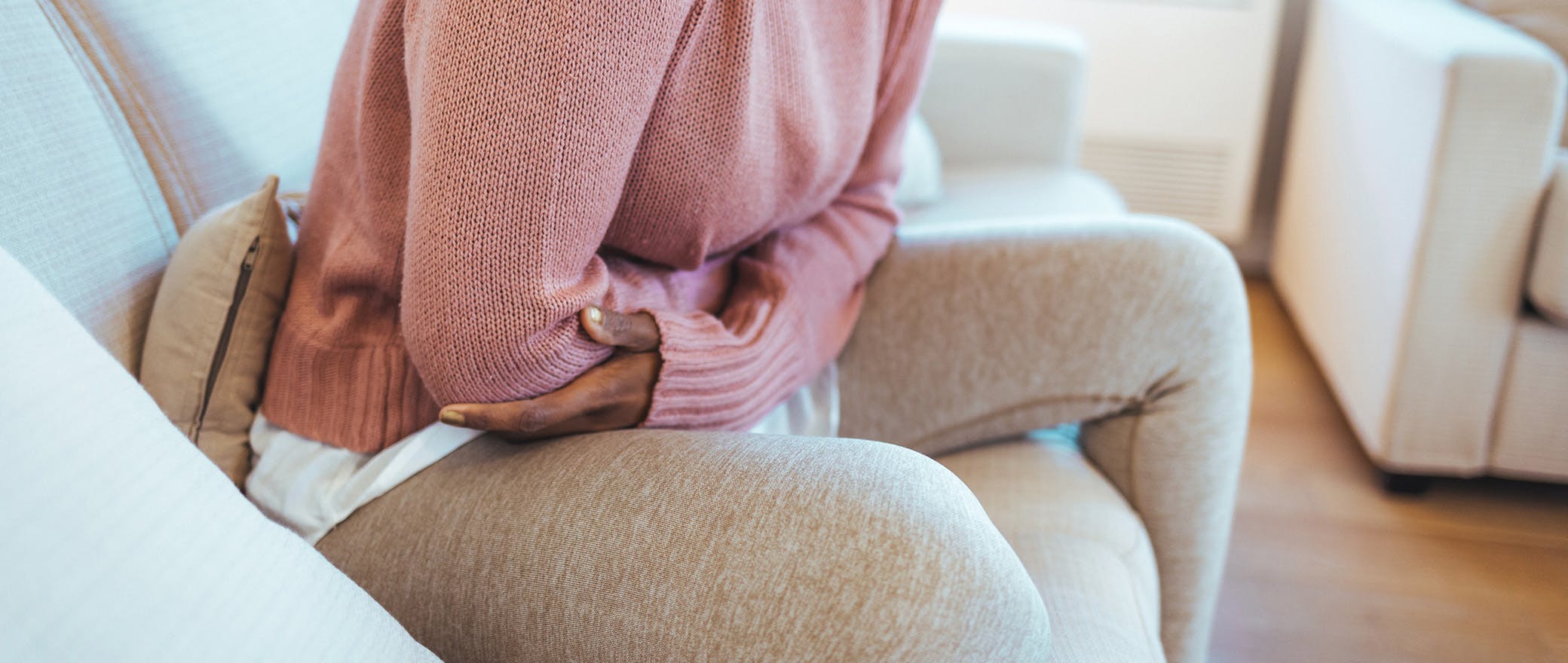 A woman is seated uncomfortably on a couch while she hugs her abdomen in pain.
