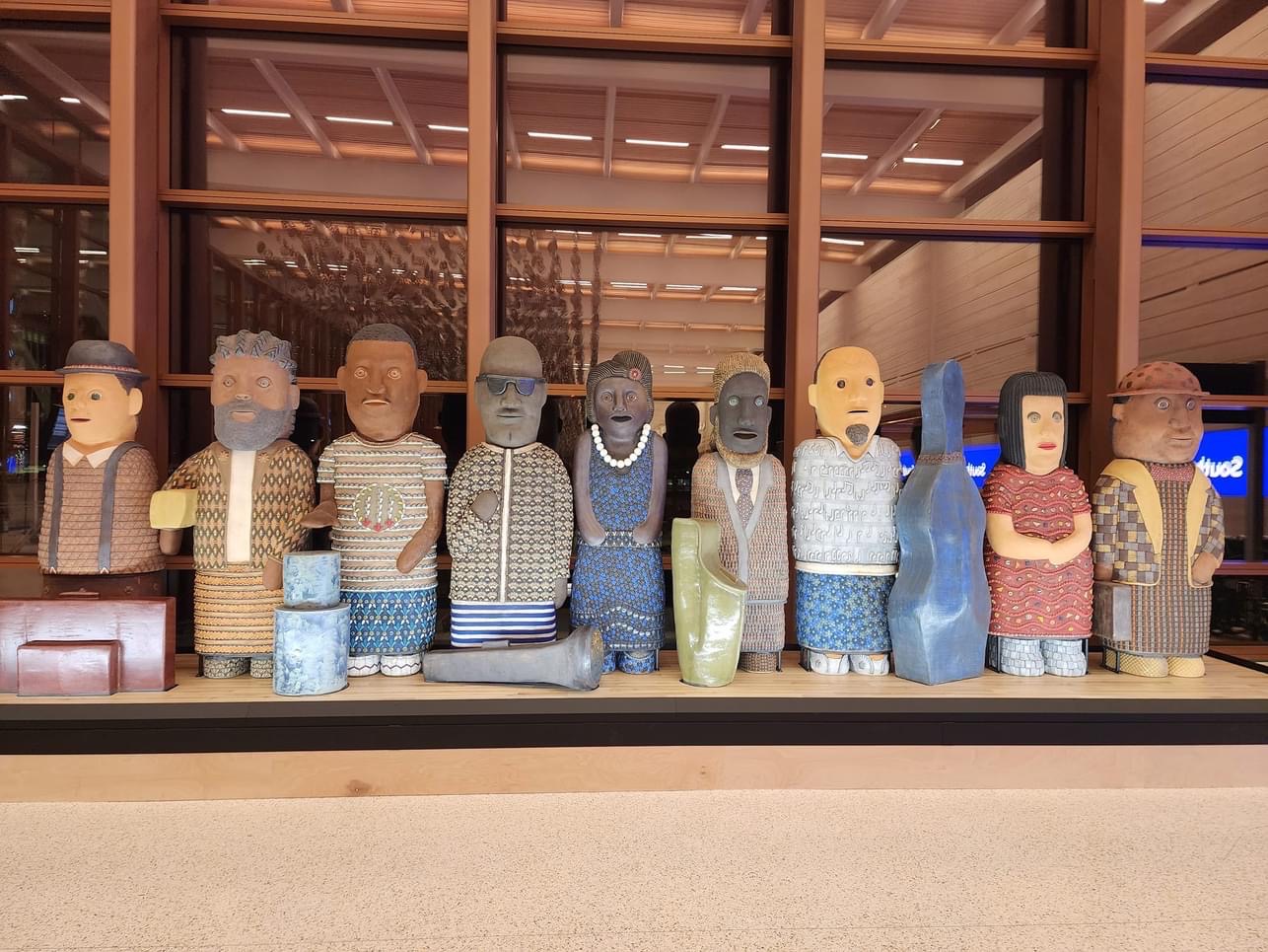 Eleven carved and painted human-like figures stand in a row, each with distinct clothing patterns, colors, and facial expressions, displayed in front of a large window
