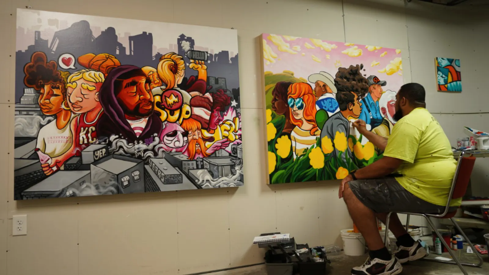 Artist JT Daniels seated in a studio painting a colorful canvas depicting diverse figures among sunflowers, with a finished panel showing a vibrant cityscape and stylized characters on the adjacent wall.