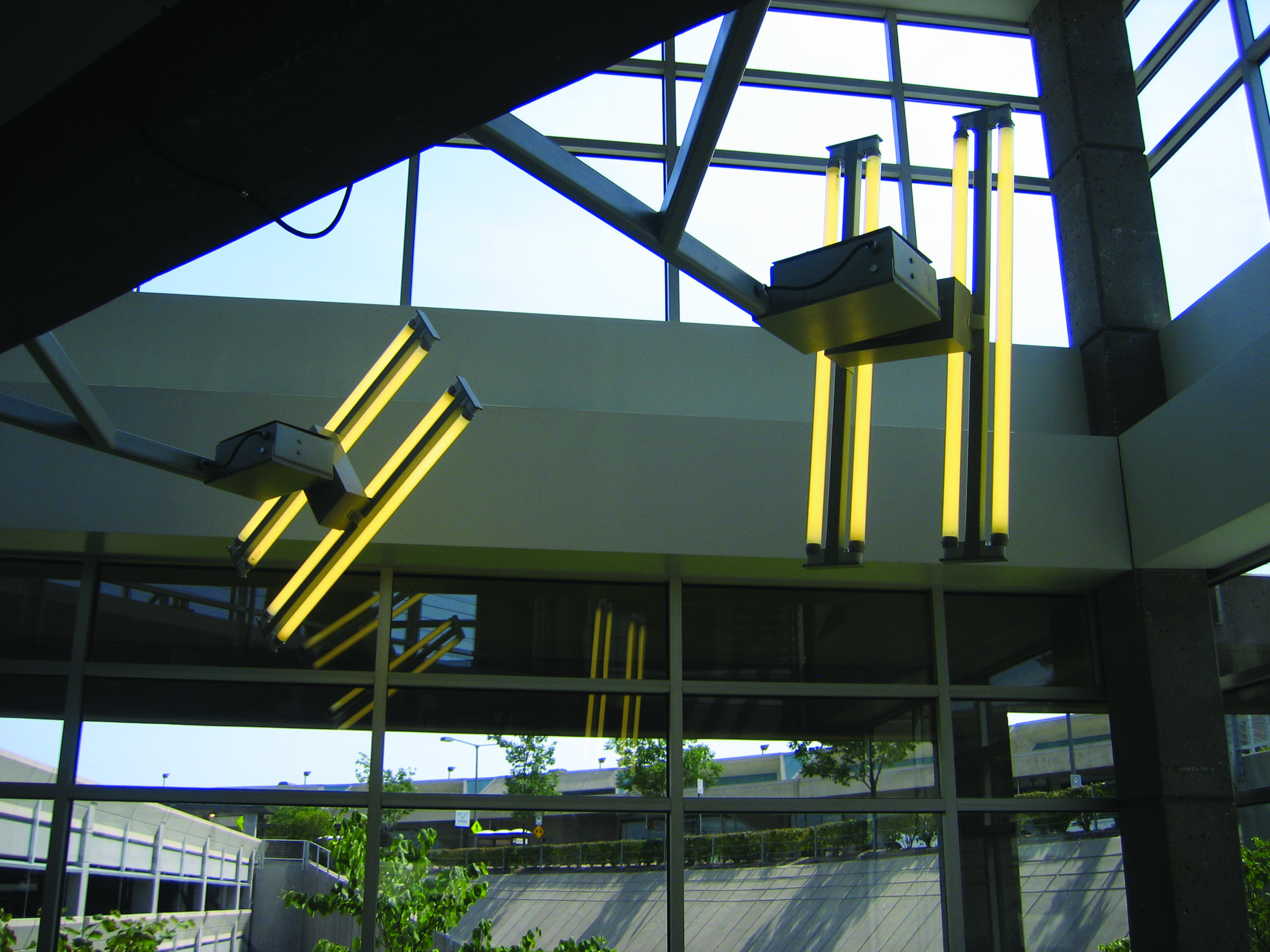 Indoor light installation featuring clusters of yellow fluorescent tubes mounted vertically and diagonally near large glass windows in a modern atrium.