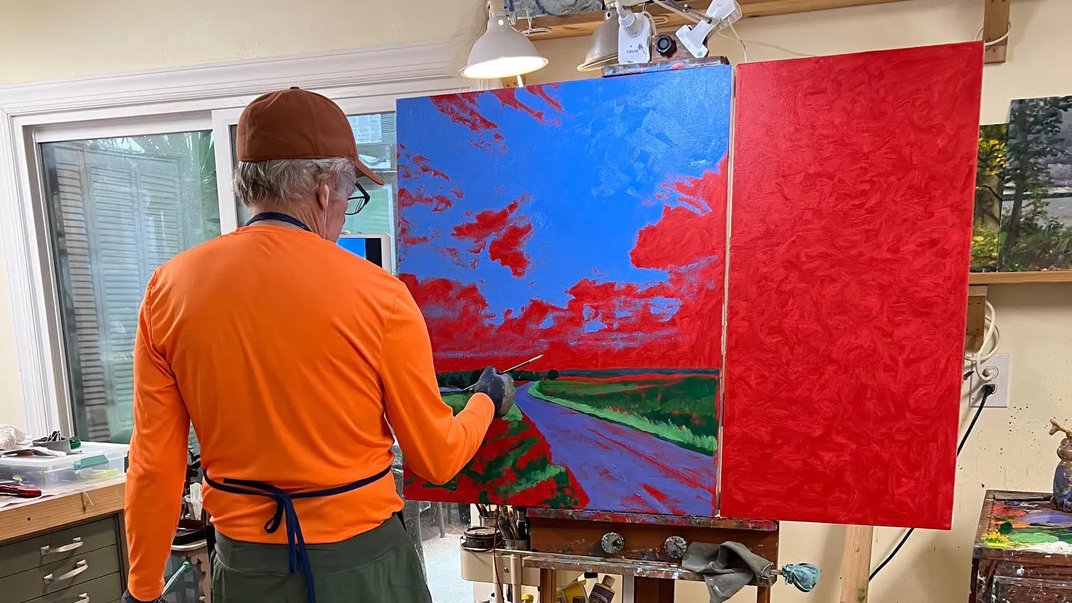 Artist John Louder painting a vibrant landscape with bold red and blue underpainting in a well-lit studio