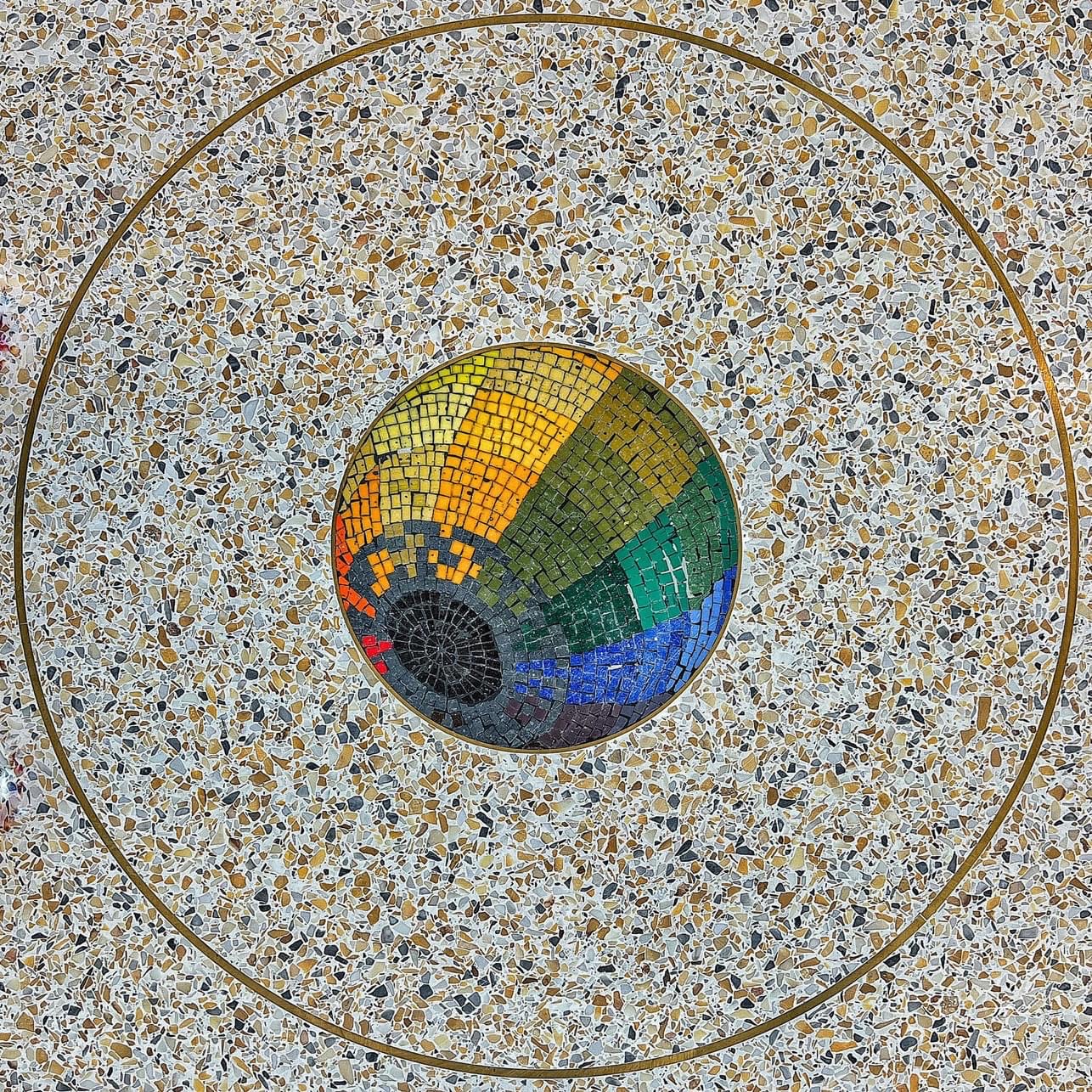 Colorful circular mosaic embedded in terrazzo flooring, featuring a black center with radiating segments in red, yellow, green, and blue, resembling a stylized iris or spectrum.