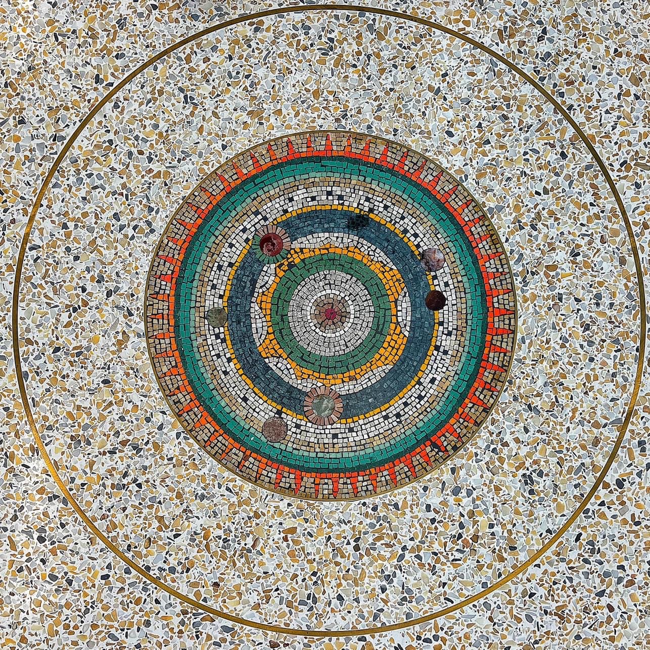 Colorful circular mosaic set in terrazzo flooring with concentric rings, planetary symbols, and radiating red triangle patterns resembling a cosmic diagram.