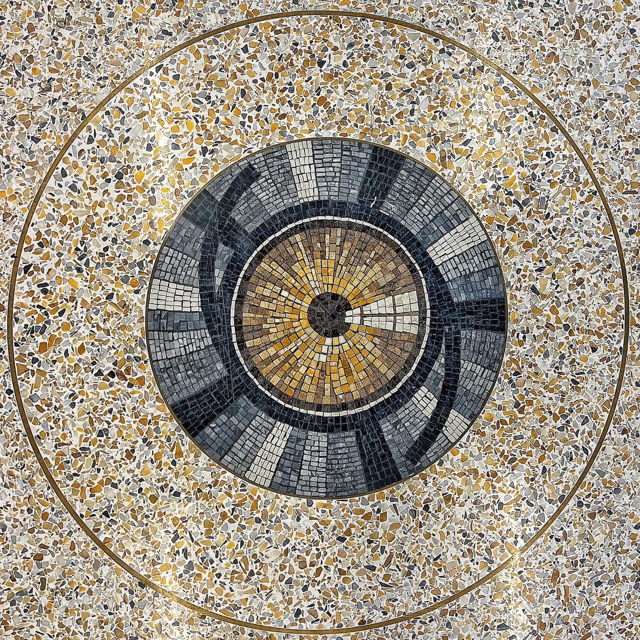 Circular floor mosaic with concentric rings and radiating patterns in black, gray, white, and gold, resembling a stylized architectural or mechanical diagram.