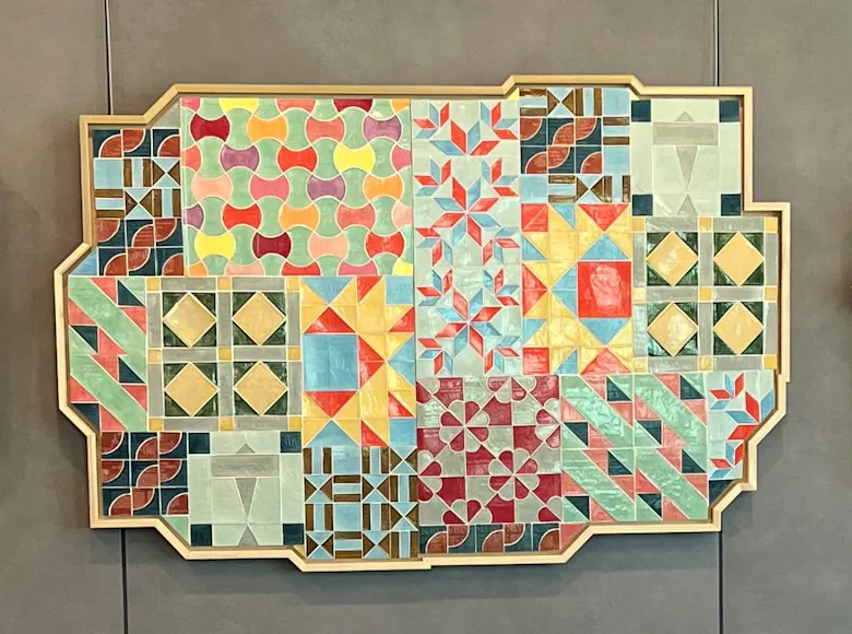 Three quilt-inspired geometric artworks in ornate frames, featuring vibrant, interlocking patterns in a variety of colors and shapes, mounted on a gray wall.