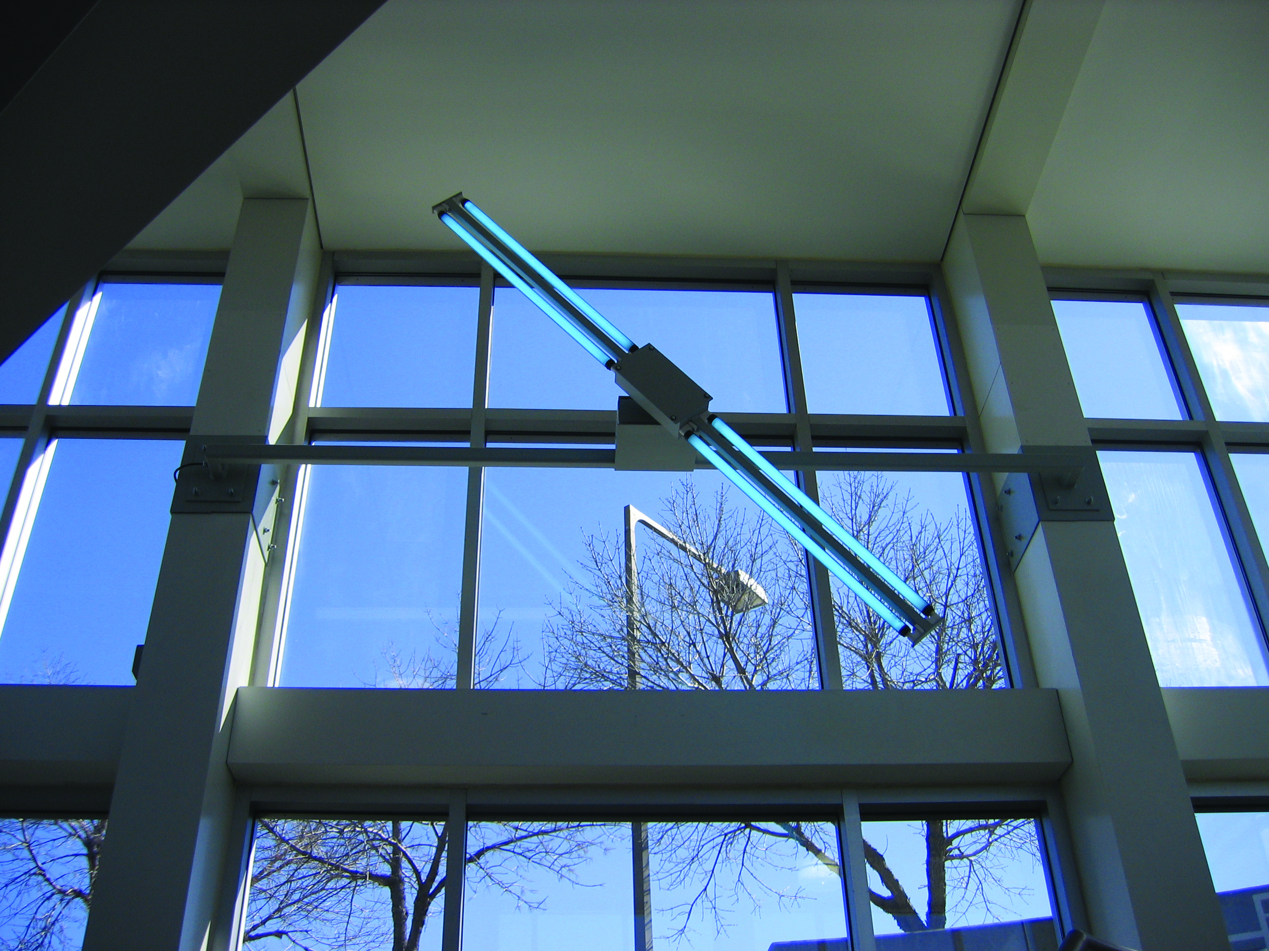 Diagonal light sculpture with glowing blue tubes mounted inside a bright airport walkway, framed by tall glass windows and outdoor trees.