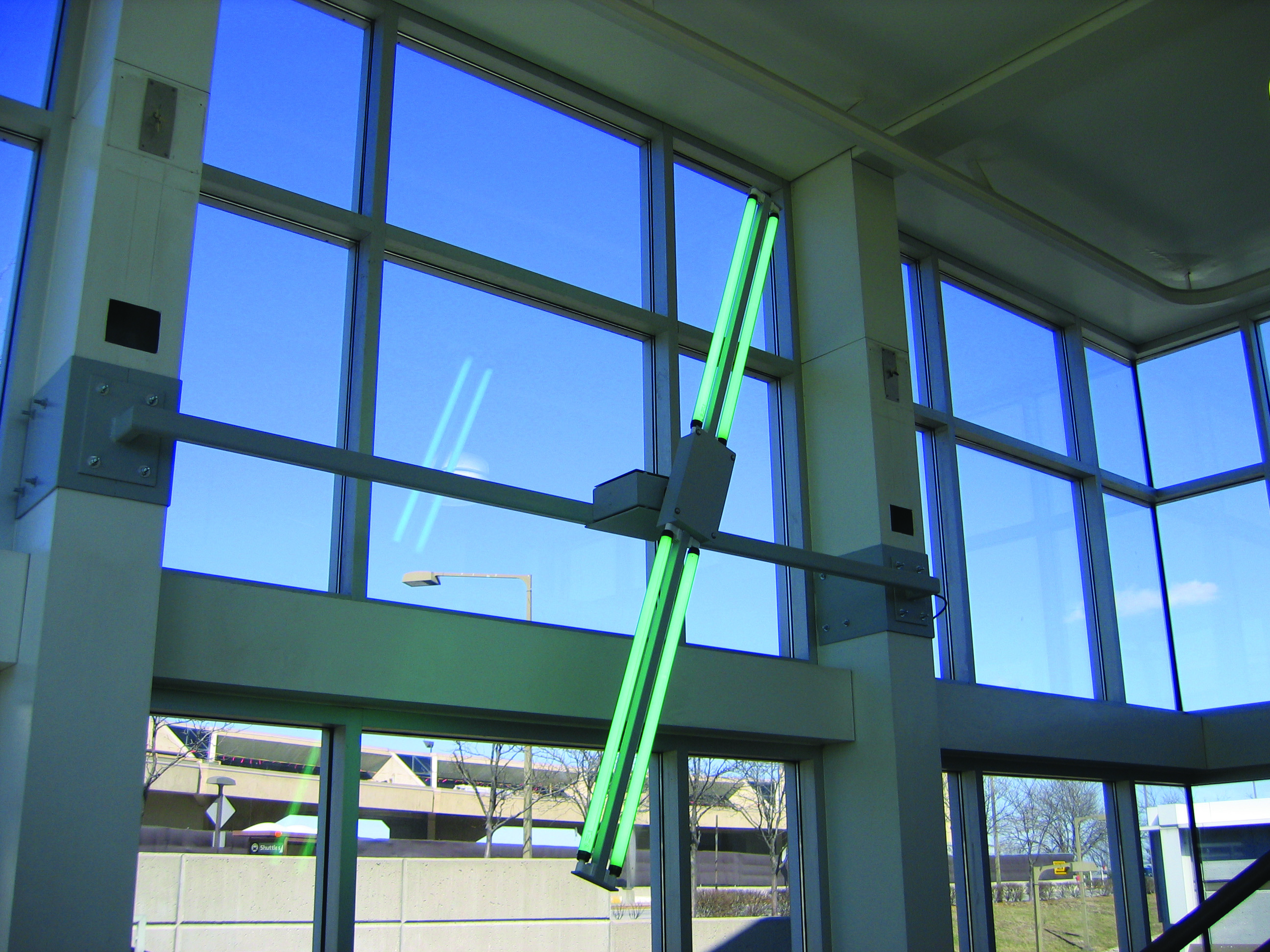 Sculptural light fixture with glowing vertical tubes mounted at an angle inside a sunlit glass-walled airport corridor.