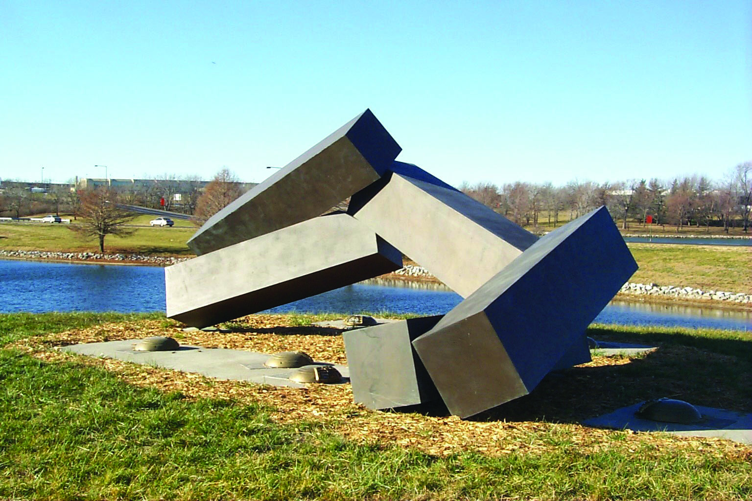 Large abstract metal sculpture of interlocking rectangular beams resting on the ground near a pond, with grassy surroundings and a clear sky.