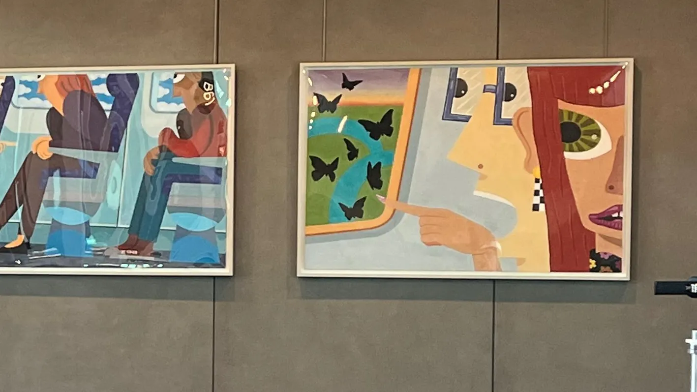 Three colorful paintings depicting stylized passengers on airplanes, each framed and mounted on a gray wall. The left shows a woman looking out a window with birds outside. The center shows multiple seated passengers. The right shows a woman pointing out a window with butterflies flying by.