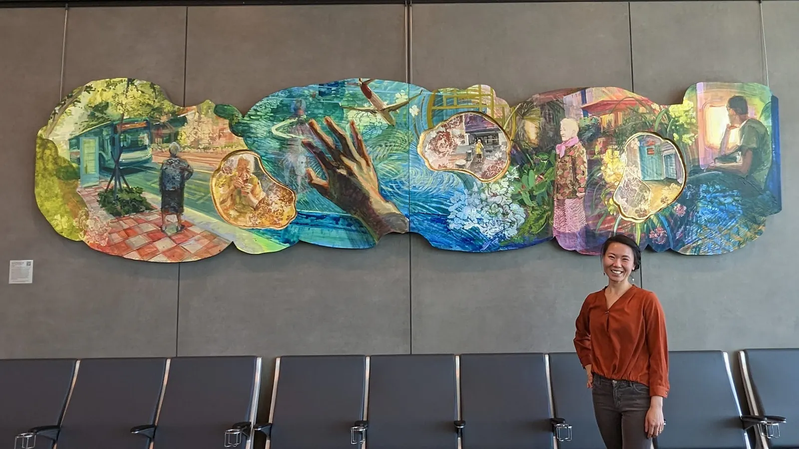 Artist Kathy Liao standing and smiling in front of a completed mural at Kansas City International Airport, depicting dreamlike vignettes of daily life and memory in vibrant color.