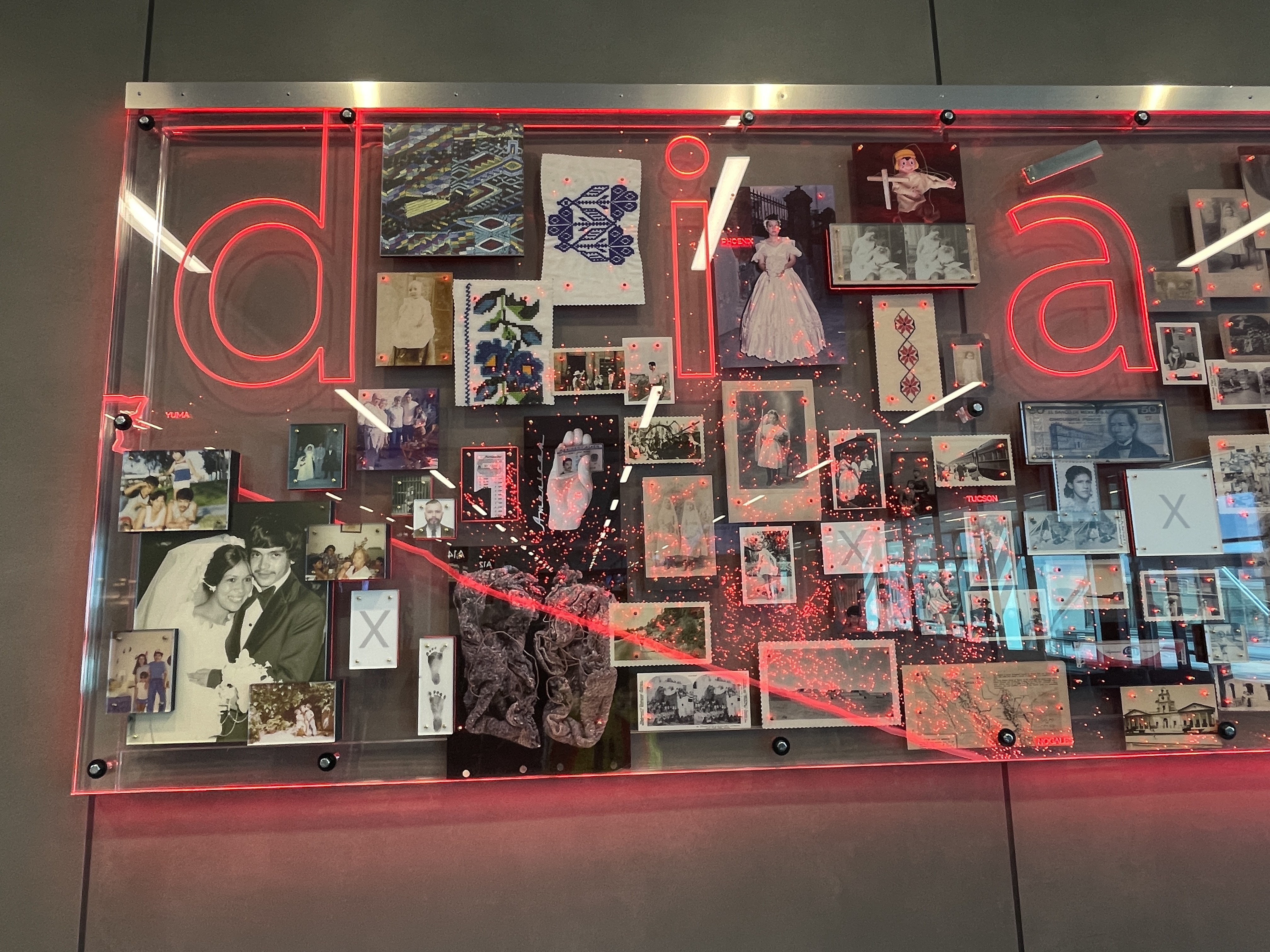 Close-up of a mixed-media collage with family photos, embroidered textiles, and cultural artifacts, overlaid with glowing red neon letters spelling “diasporia.”