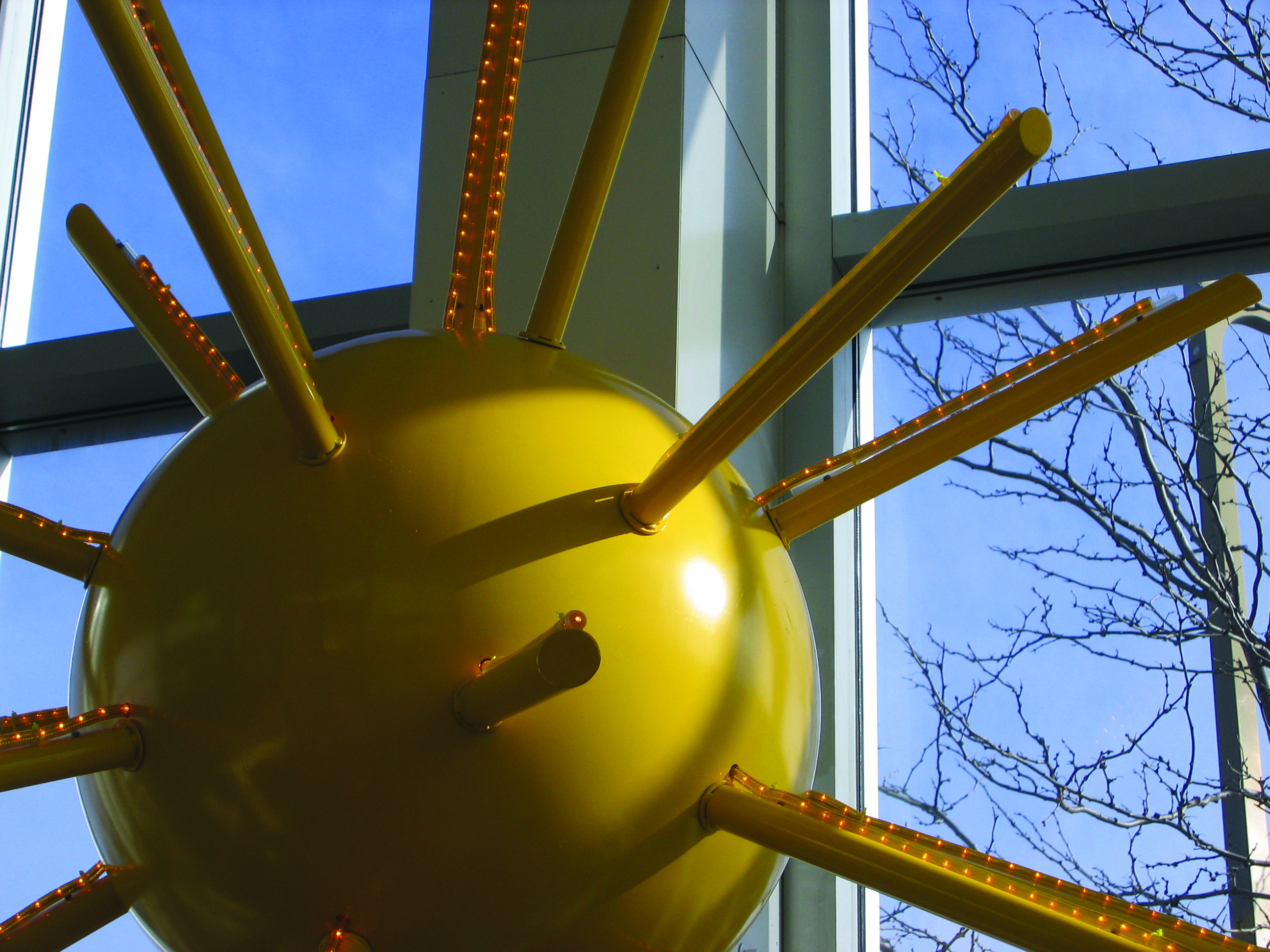 Close-up of a glossy yellow sunburst sculpture with radiating rods lit by small bulbs, positioned inside a glass-walled space with blue sky and tree branches visible outside.