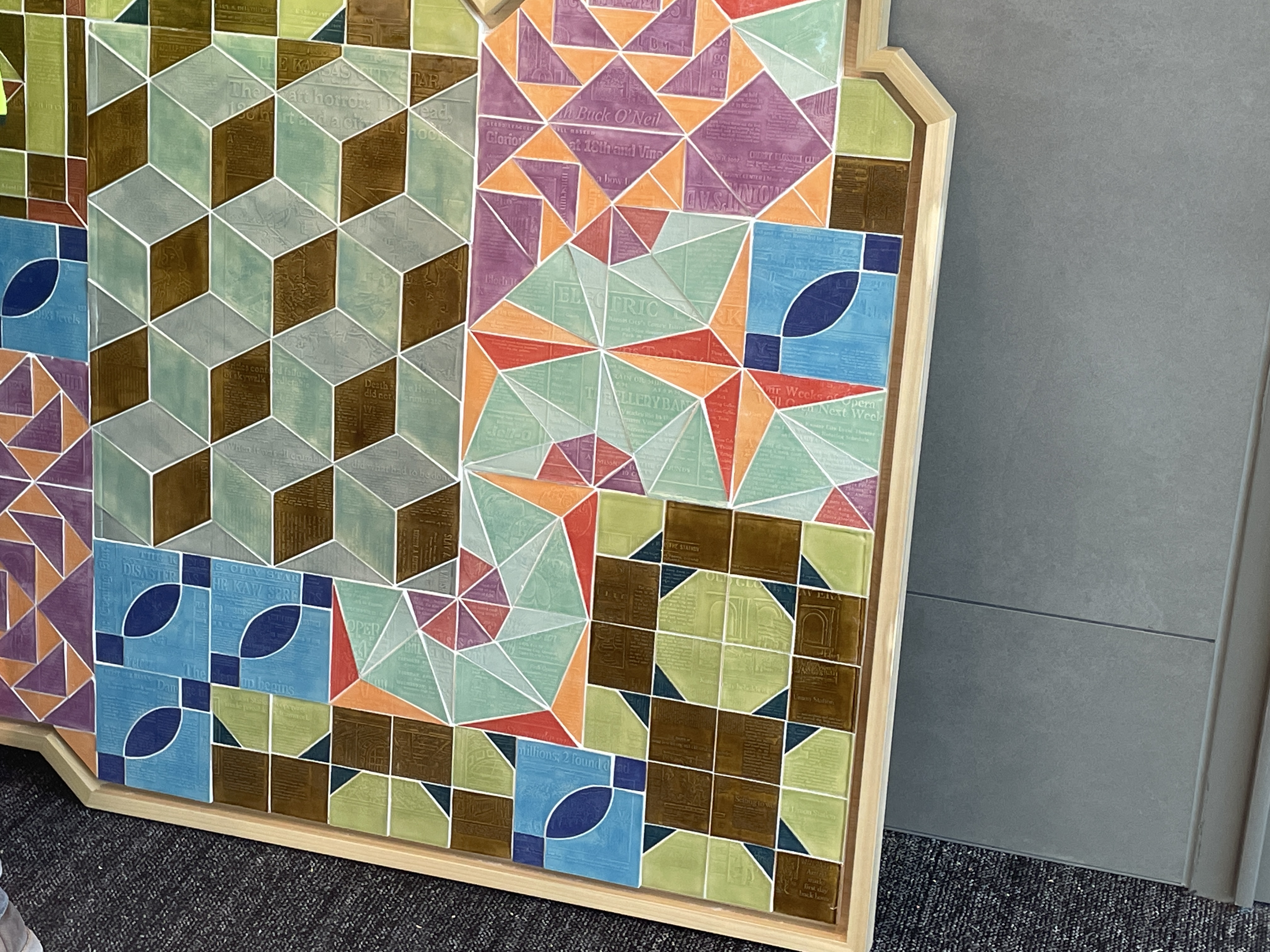 Detail view of a geometric artwork composed of colorful triangular, square, and cube-like patterns, some overlaid with faint printed text, all framed within a custom-cut wooden edge.