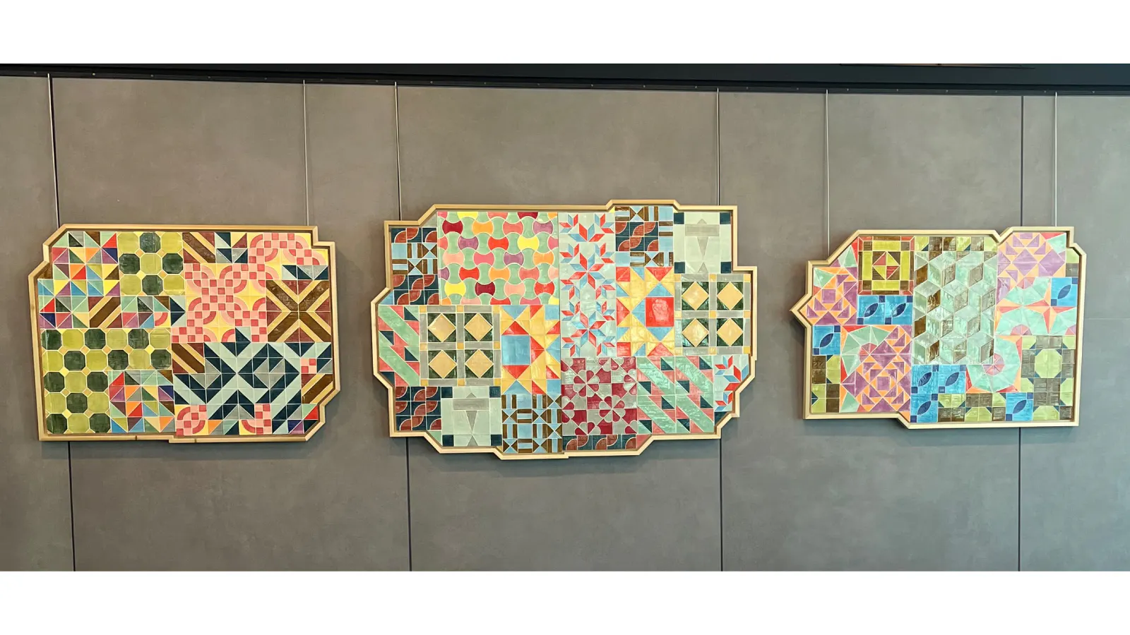 Three quilt-inspired geometric artworks in ornate frames, featuring vibrant, interlocking patterns in a variety of colors and shapes, mounted on a gray wall.