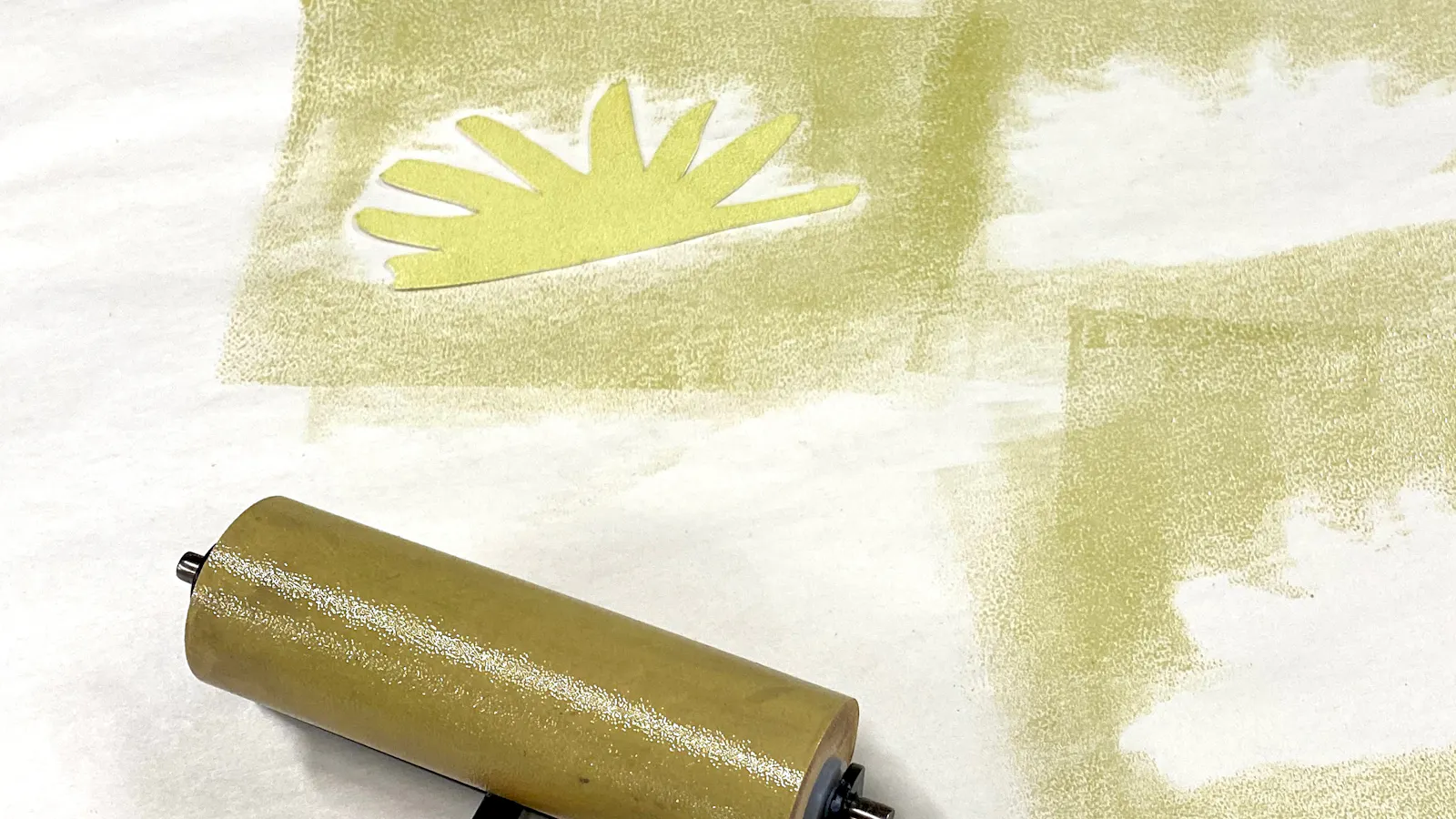 Close-up of a stencil printing process showing a brayer roller and several leafy, fan-shaped forms in light green and brown being applied to a white surface.