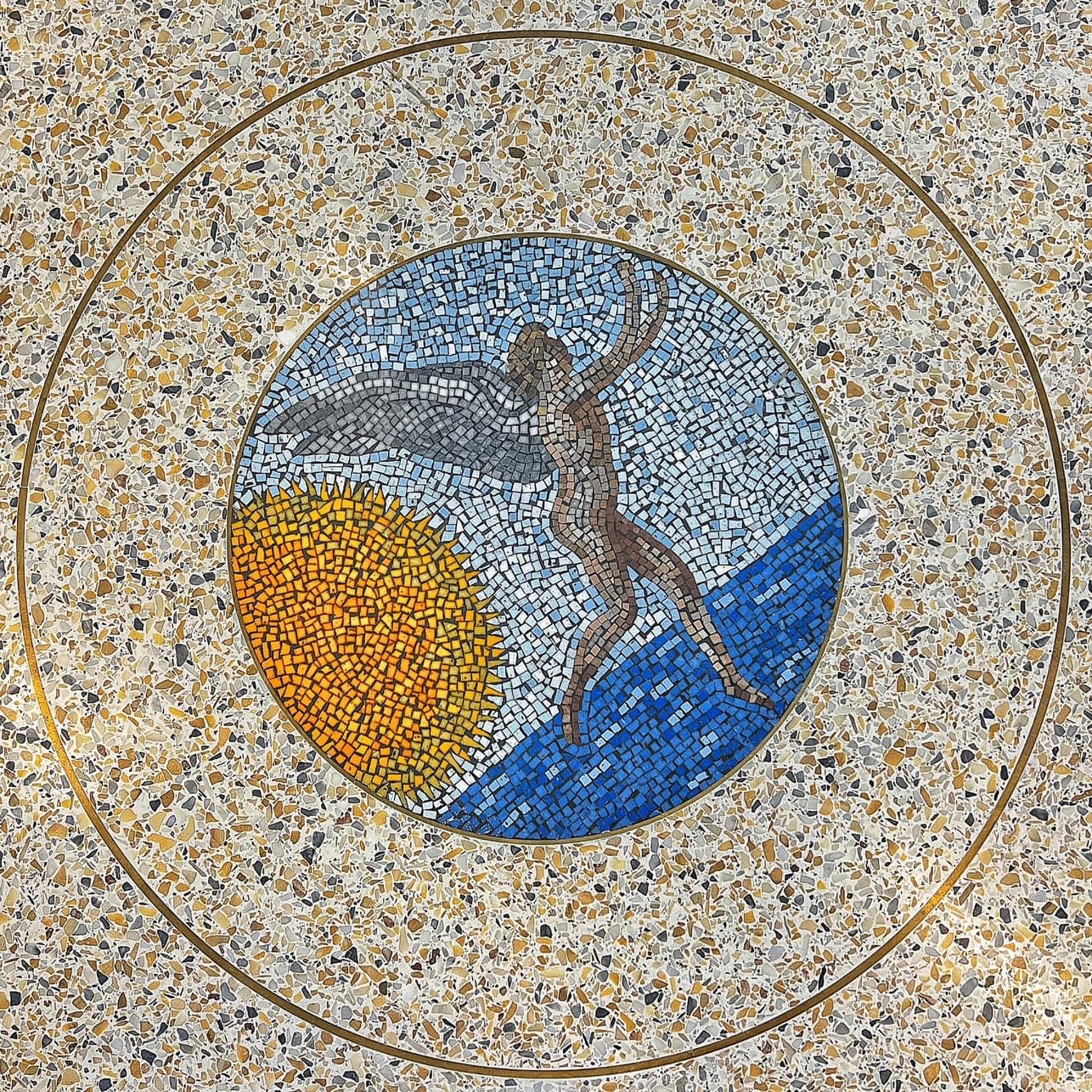 Mosaic in a terrazzo floor depicting a winged human figure reaching upward beside a glowing sun over a blue horizon, enclosed by a brass ring.