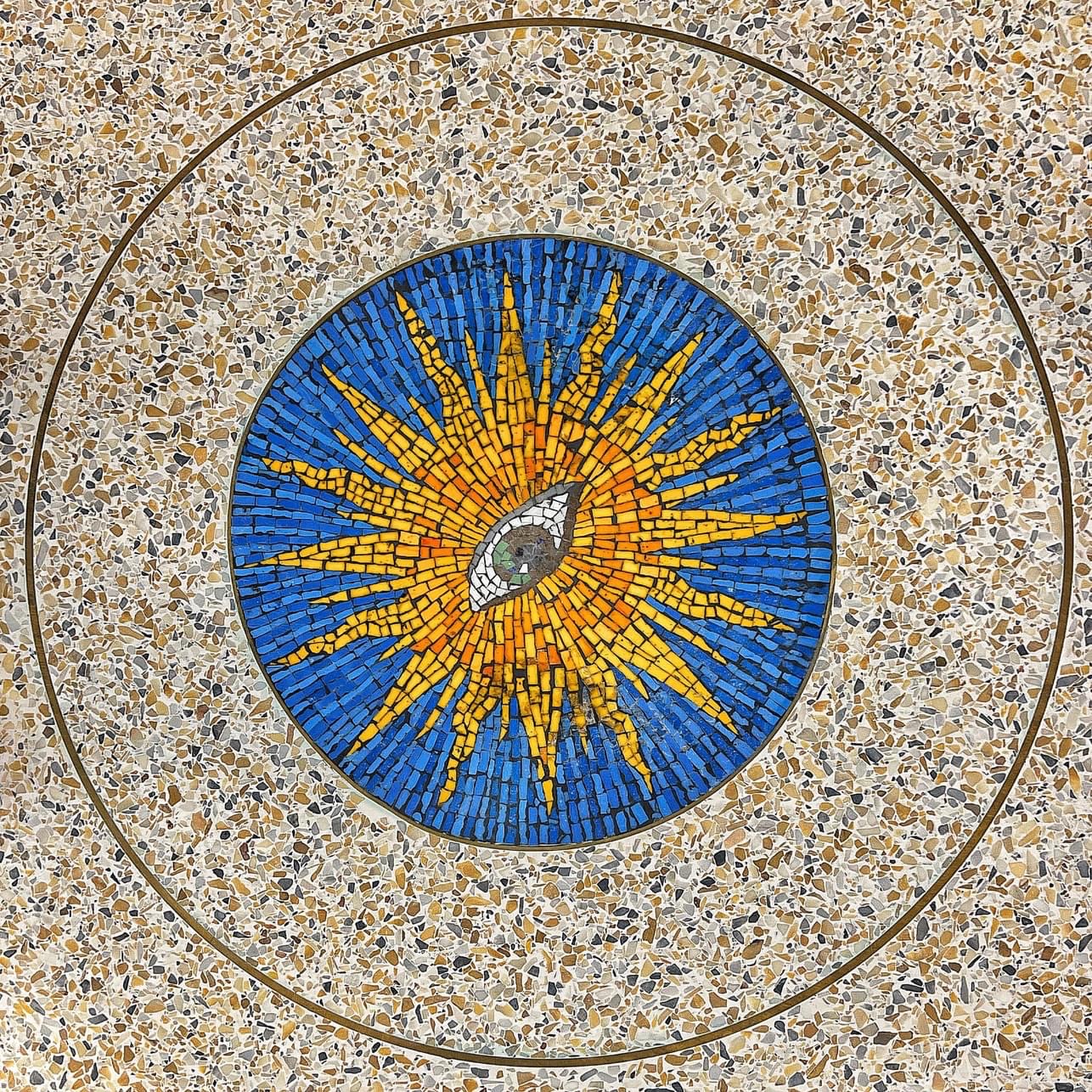 Circular mosaic in a terrazzo floor depicting a central eye surrounded by yellow rays against a blue background, resembling a radiant sun.