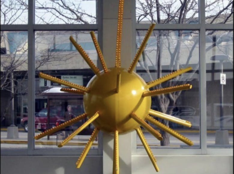 Yellow sun-shaped sculpture with radiating rods mounted on a pillar inside a glass-walled space, with street and pedestrian visible outside.