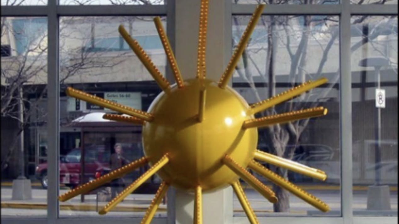 Yellow sun-shaped sculpture with radiating rods mounted on a pillar inside a glass-walled space, with street and pedestrian visible outside.