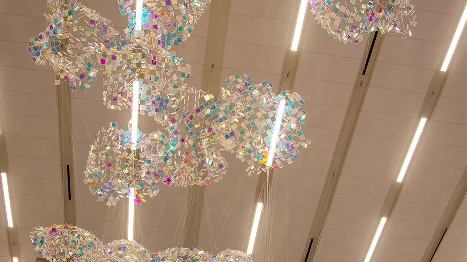 Looking up at a cascading sculpture of shimmering, iridescent mesh suspended beneath ceiling lights at Kansas City International Airport.