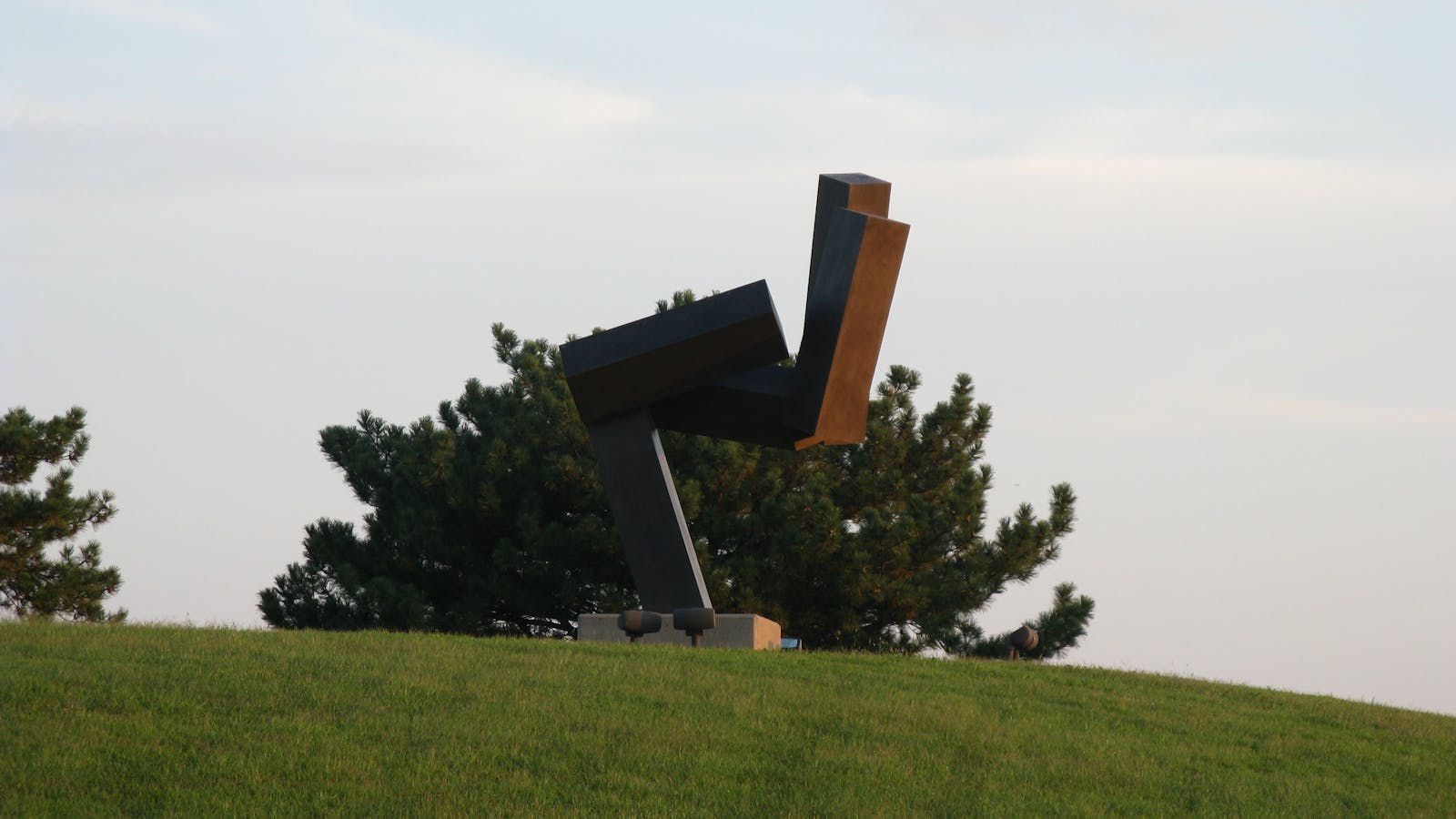 Abstract metal sculpture of angled beams mounted on a hilltop, silhouetted against trees and a soft evening sky.