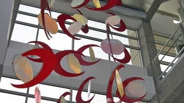 Tall hanging mobile in a glass atrium featuring red crescent forms and textured oval panels in white, yellow, and pink, suspended in a swirling vertical arrangement.